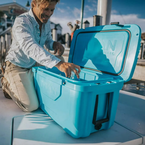 Man opening a teal cooler box on a dock with people and boats in the background