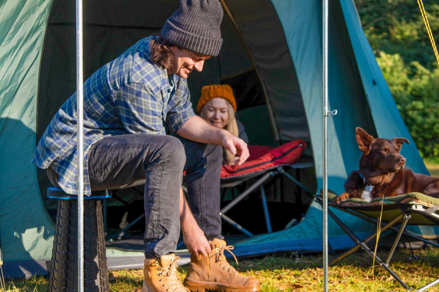 Two people and a dog sitting outside a tent in a camping setting.